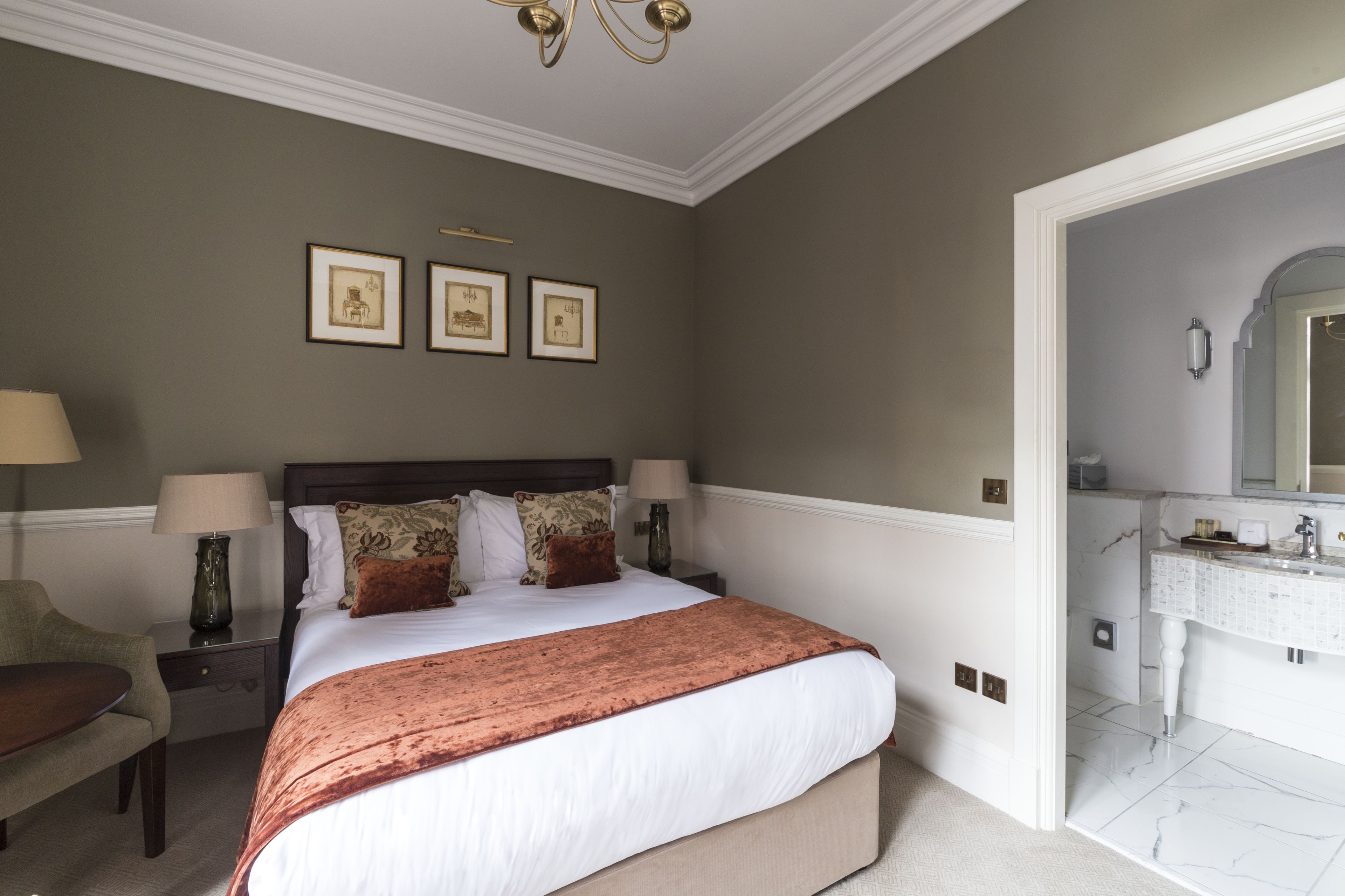A neatly arranged bedroom with a large bed, bedside tables with lamps, a chair, and a bathroom with a mirror and sink.