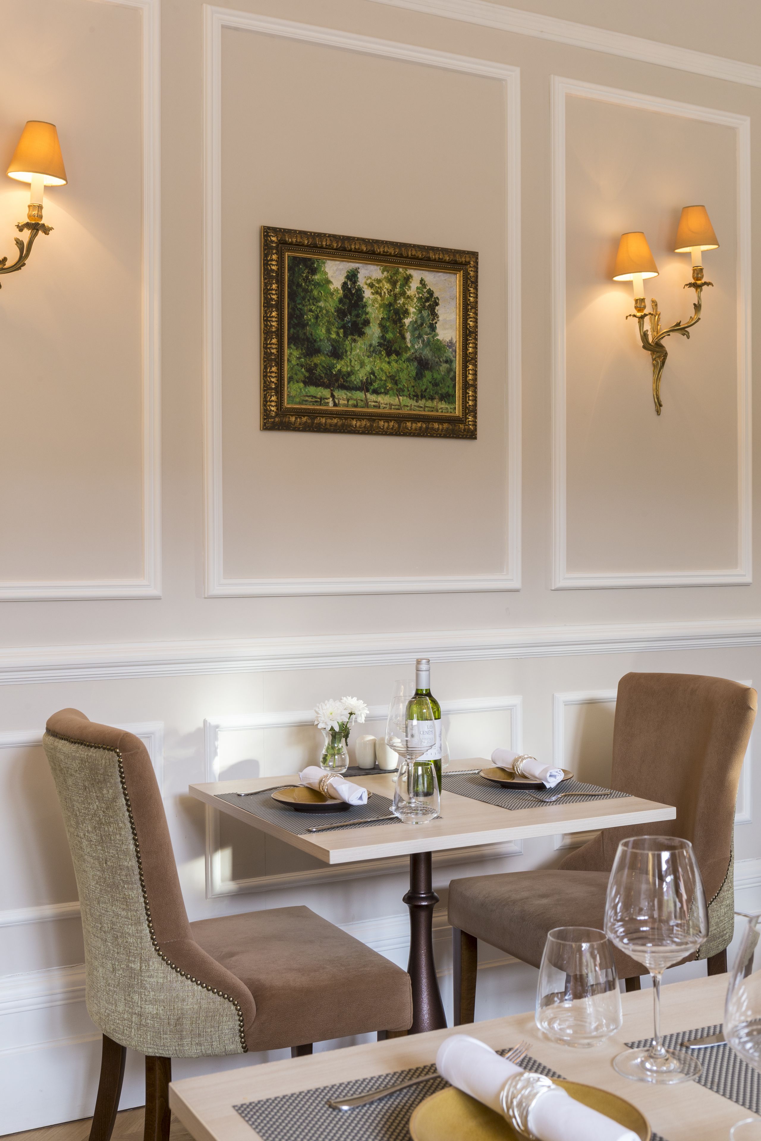 A dining area with a table set for two, featuring a painting on the wall, wall-mounted lamps, and a bottle of wine.