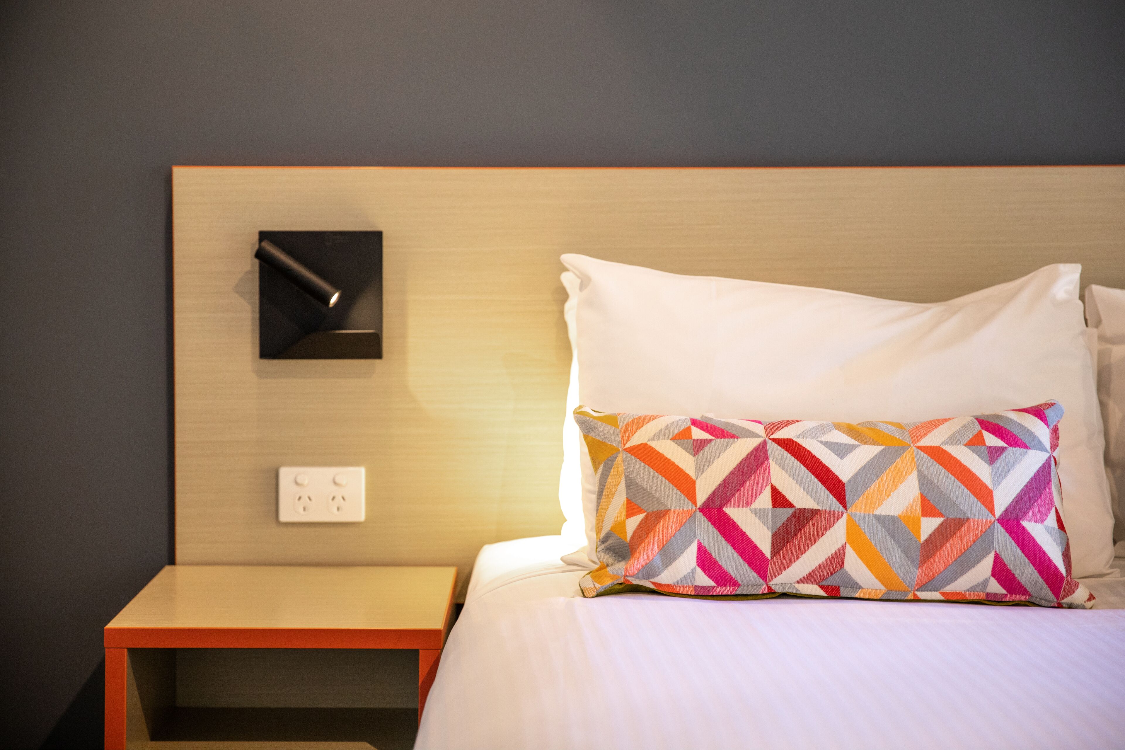 A neatly made bed with a colorful geometric pillow, a bedside table, and a wall-mounted light fixture.