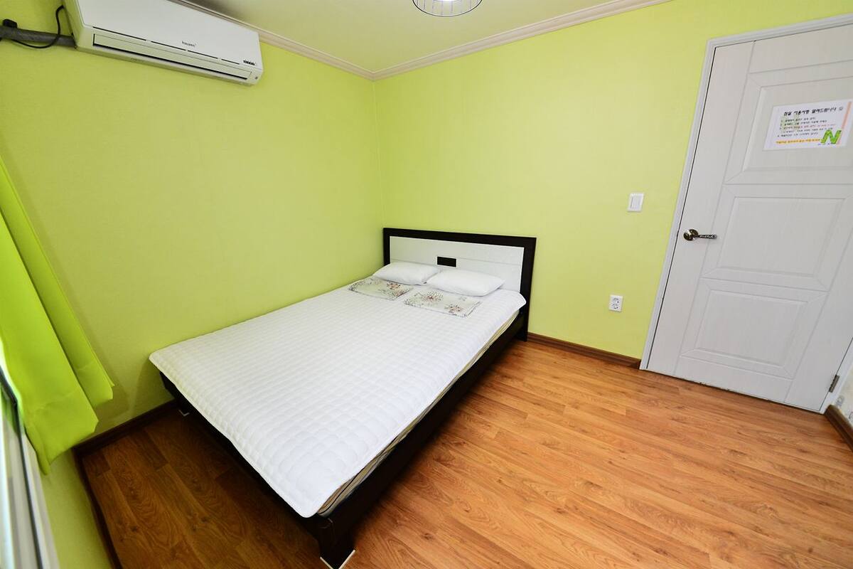 A small, single-bed room with a wooden floor, a white bed, and a wall-mounted air conditioner.