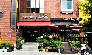 A brick building with a sign for 'Guest House NamU' and a cafe. There are potted plants and umbrellas outside.