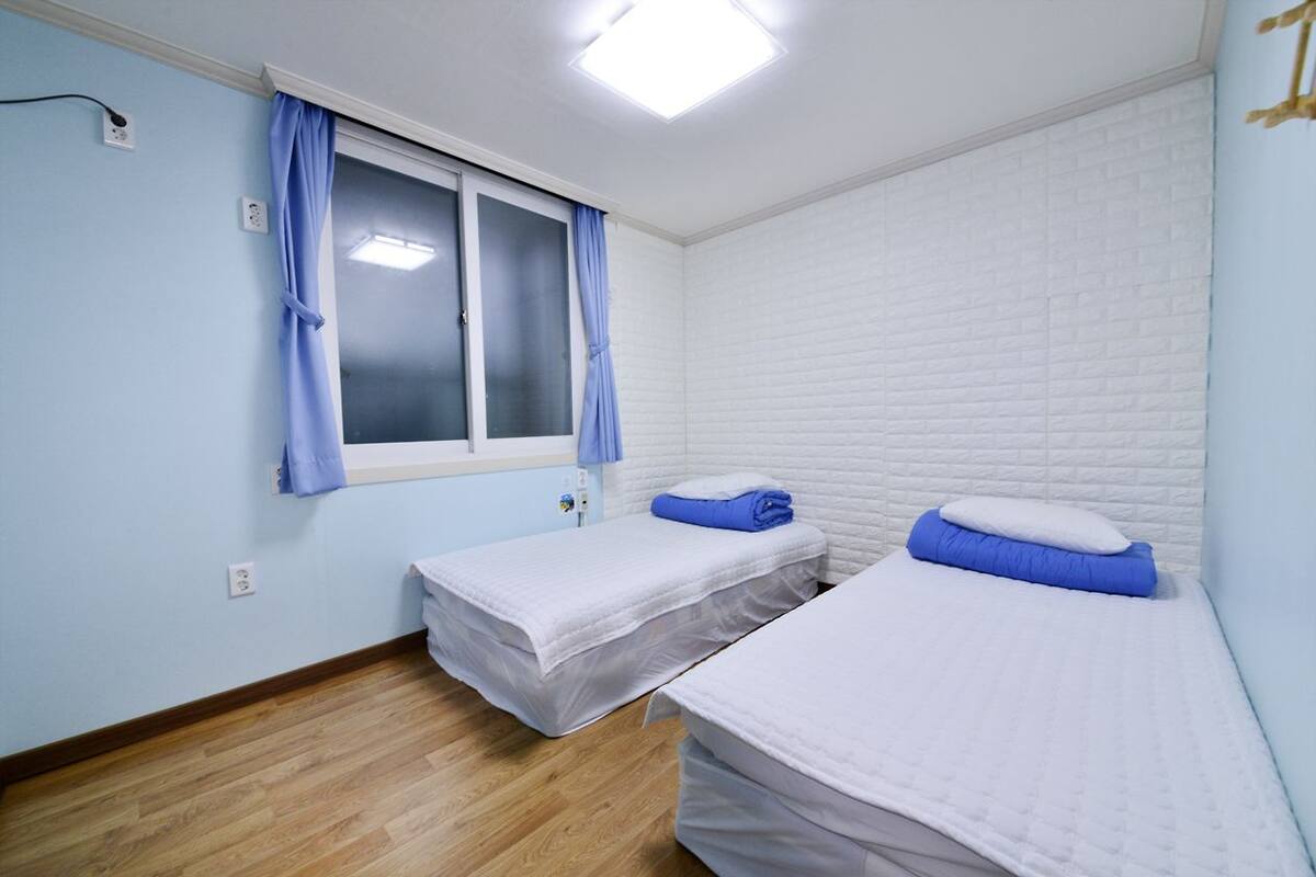 A room with two beds, a window with curtains, a white brick wall, and a wooden floor.