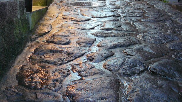 Una estrecha calle adoquinada flanqueada por edificios tradicionales con señalización en un alfabeto desconocido, lo que sugiere un área histórica o cultural.