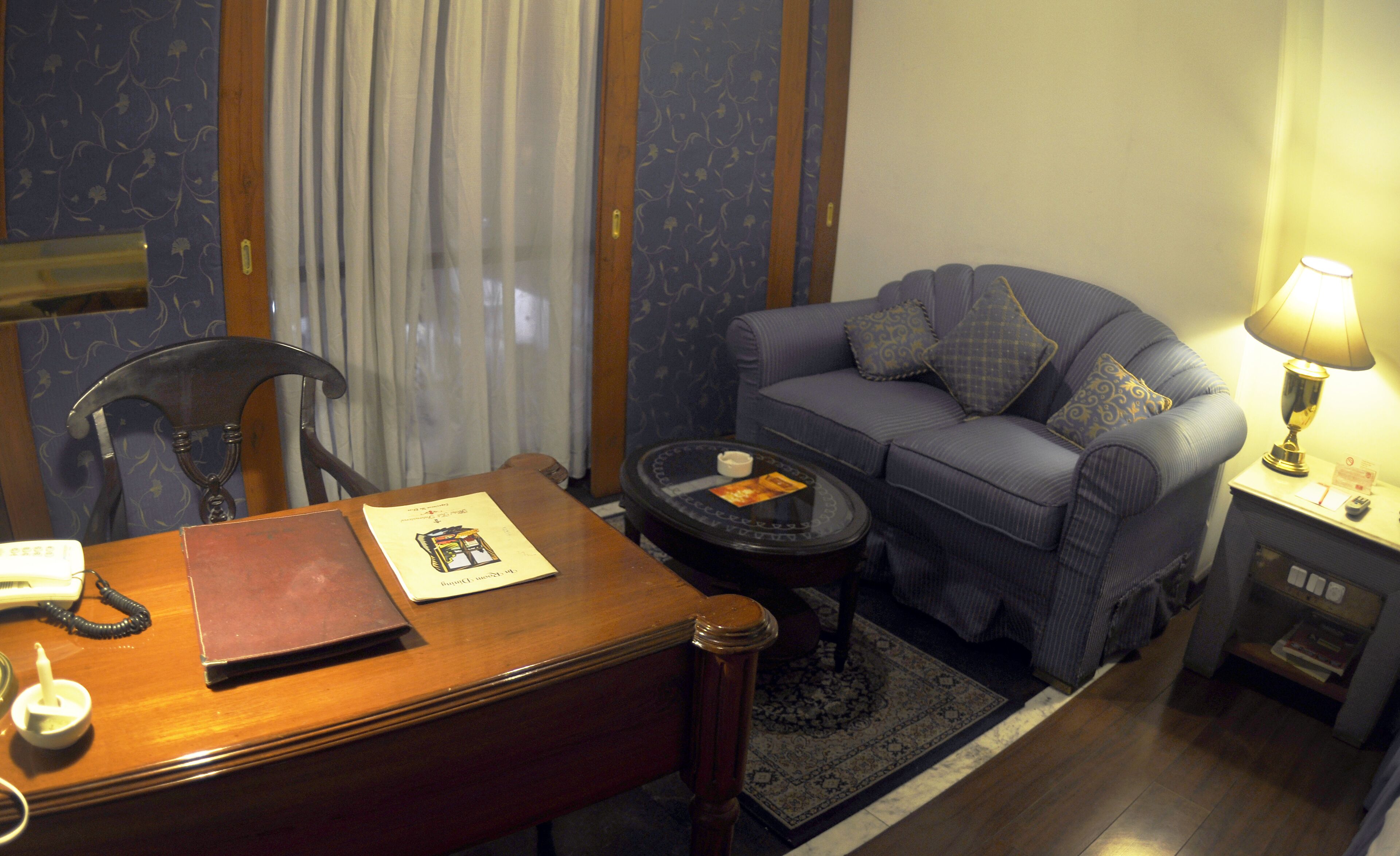 Une chambre d’hôtel avec un bureau en bois, un canapé bleu, une petite table ronde et une lampe.