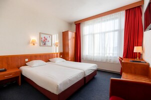 Ein Hotelzimmer mit zwei Betten, einem Holzschrank, einem Schreibtisch mit Lampe und einem großen Fenster mit roten Vorhängen.