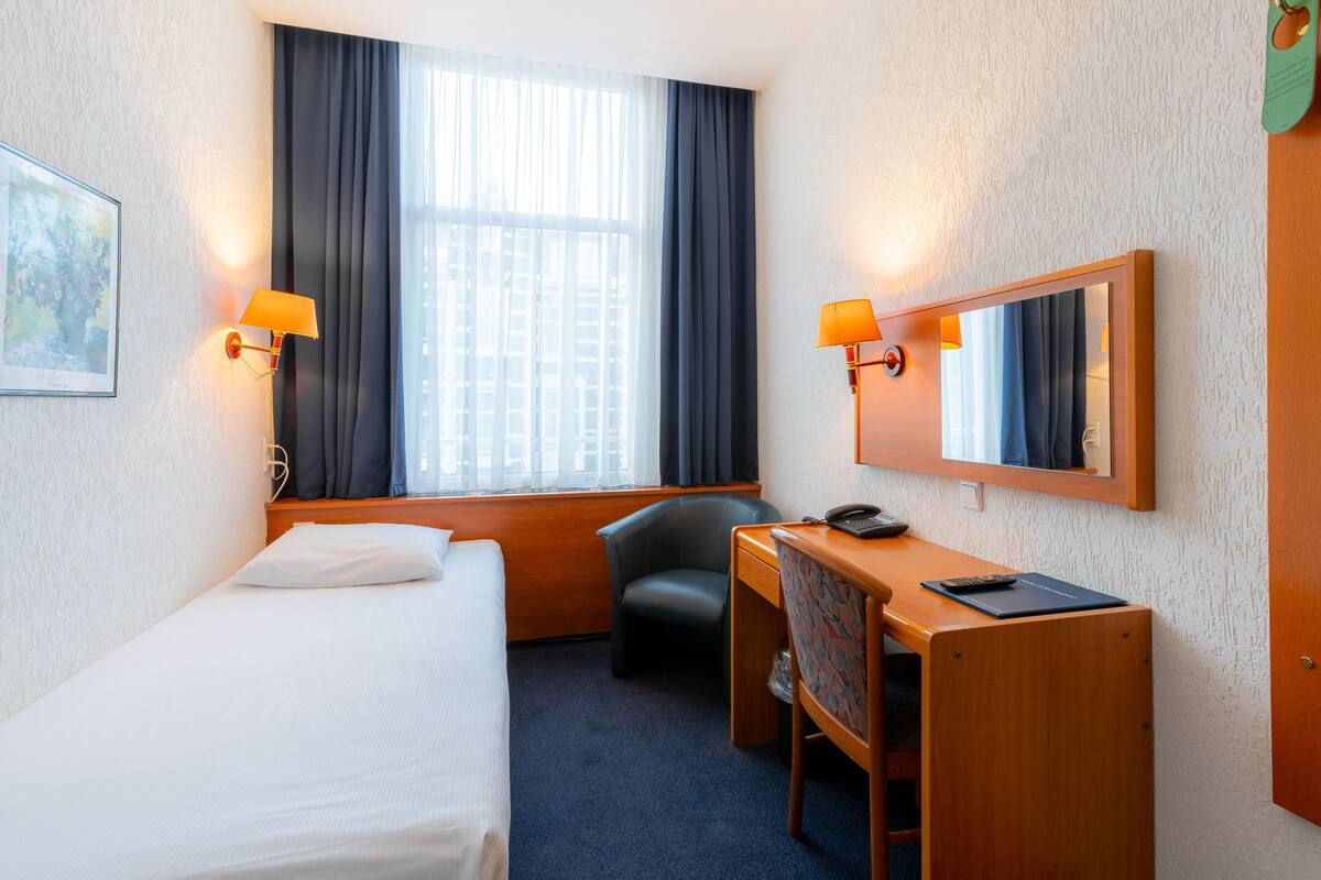 A hotel room with a bed, a desk, a chair, a desk lamp, a telephone, and a mirror.