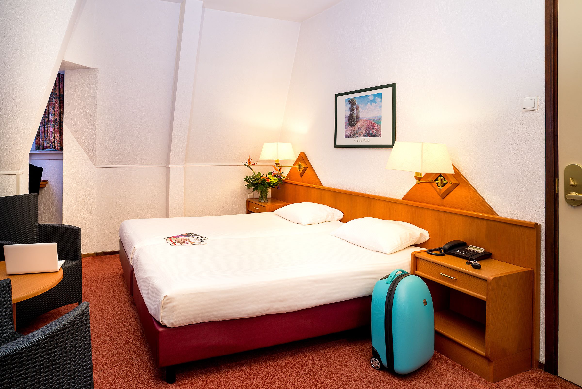 A hotel room with a bed, bedside lamps, a desk with a laptop, a suitcase, and a framed picture on the wall.