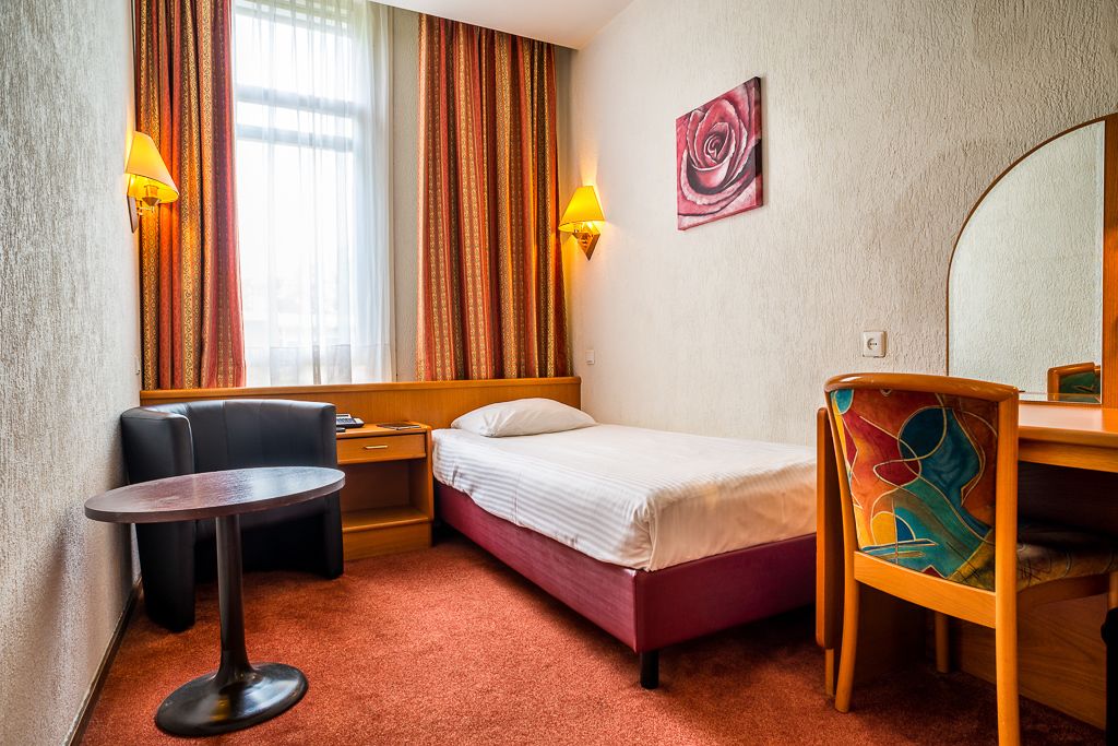 A hotel room with a bed, a desk, a chair, a small table, and a window with curtains.