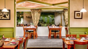 A dining area with wooden tables and chairs, a green wall, and a view of a garden through a window. - Owl Hotel (Amsterdam)
