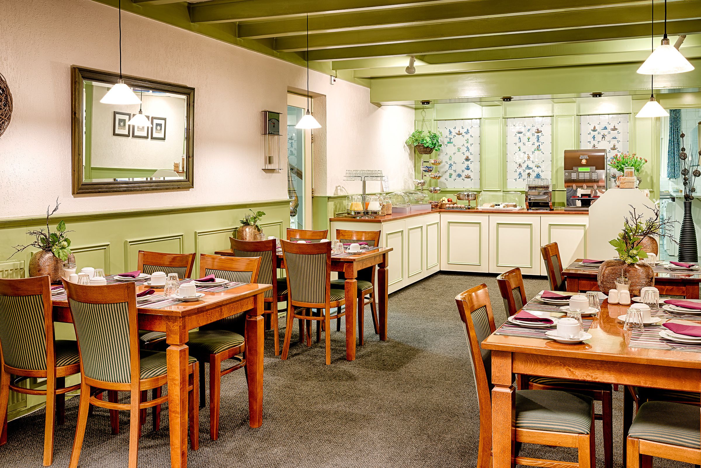 A dining area with wooden tables and chairs, a kitchen in the background, and a framed picture on the wall.