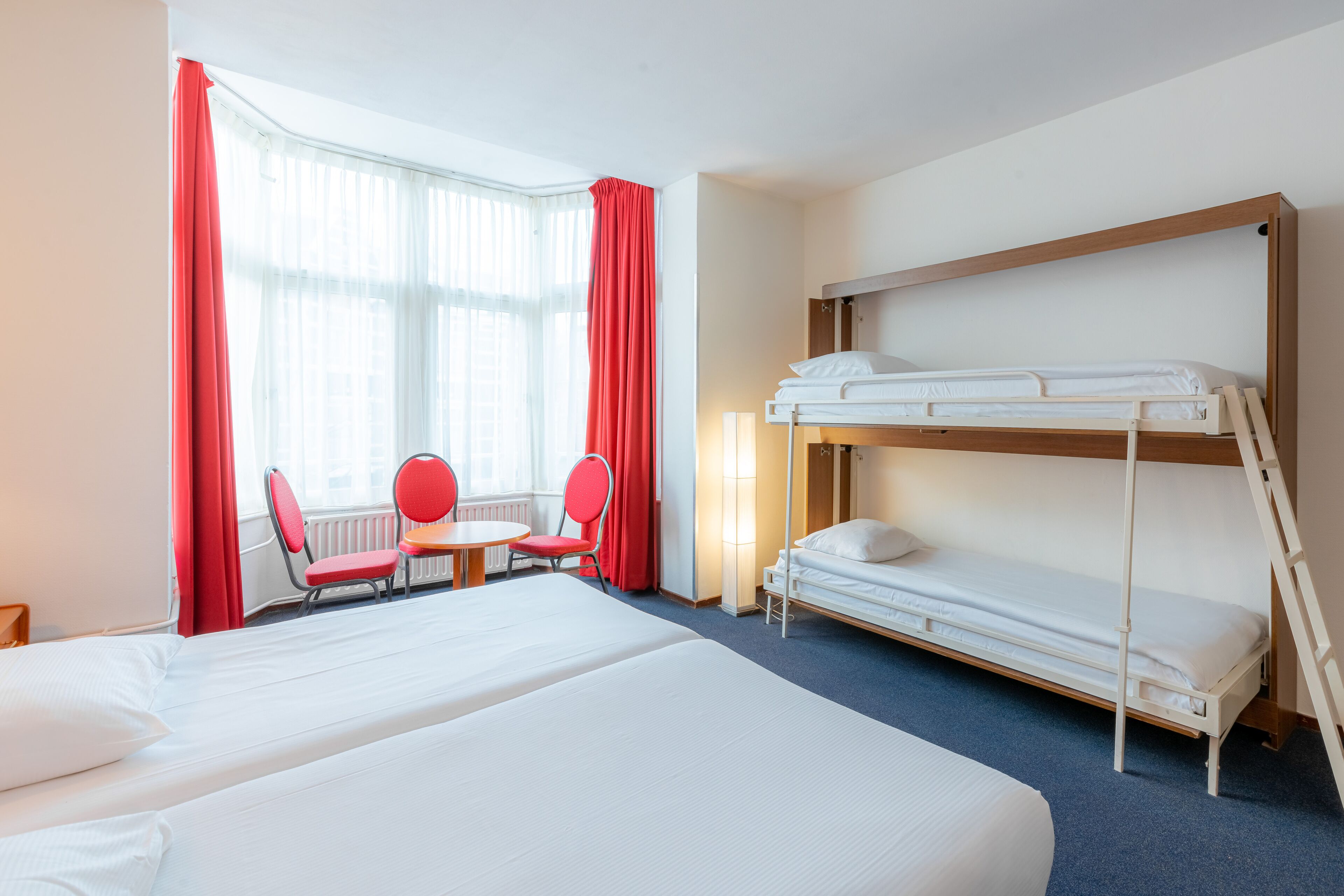 Een hotelkamer met twee bedden, een raam met rode gordijnen, een tafel met twee stoelen en een lamp.