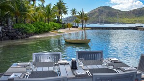 A dining table set with chairs, overlooking a calm body of water with boats and a beach in the background. - South Point Antigua Ocean Suites (English Harbour)