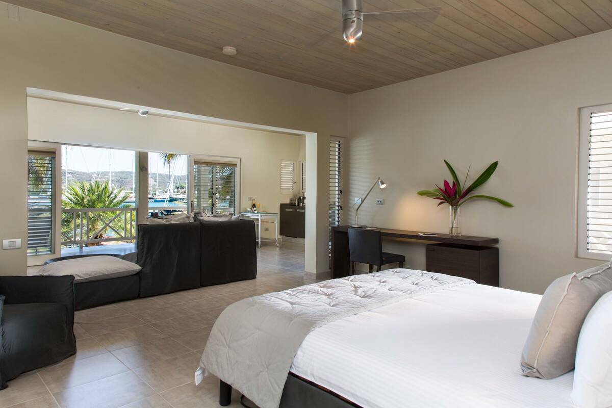 A bedroom with a large bed, a desk, a chair, and a view of the outside.