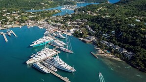 An aerial view of a marina with several yachts docked, surrounded by lush greenery and residential buildings. - South Point Antigua Ocean Suites (English Harbour)
