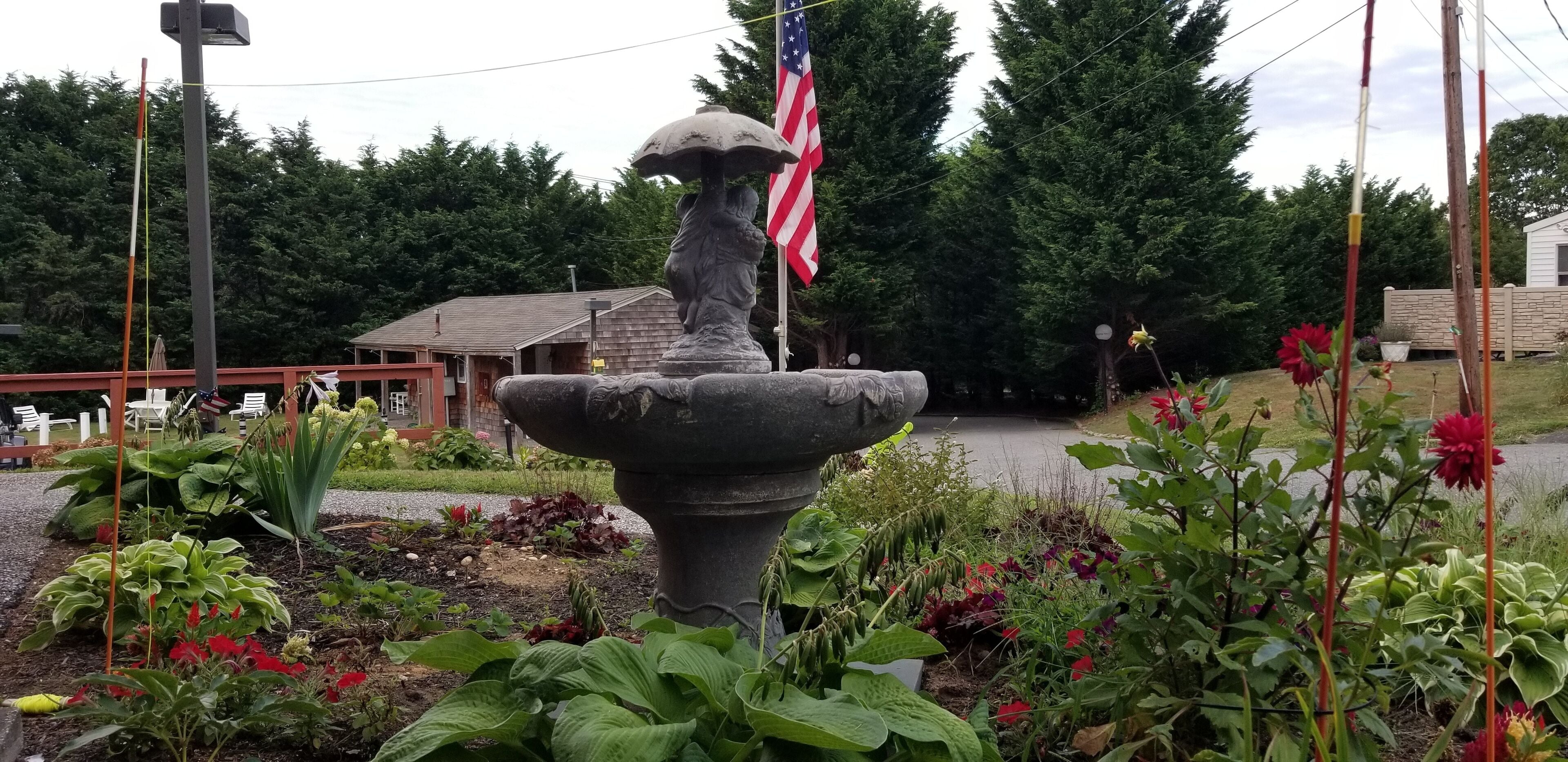 Un jardín con una fuente de piedra, una bandera estadounidense y diversas plantas.