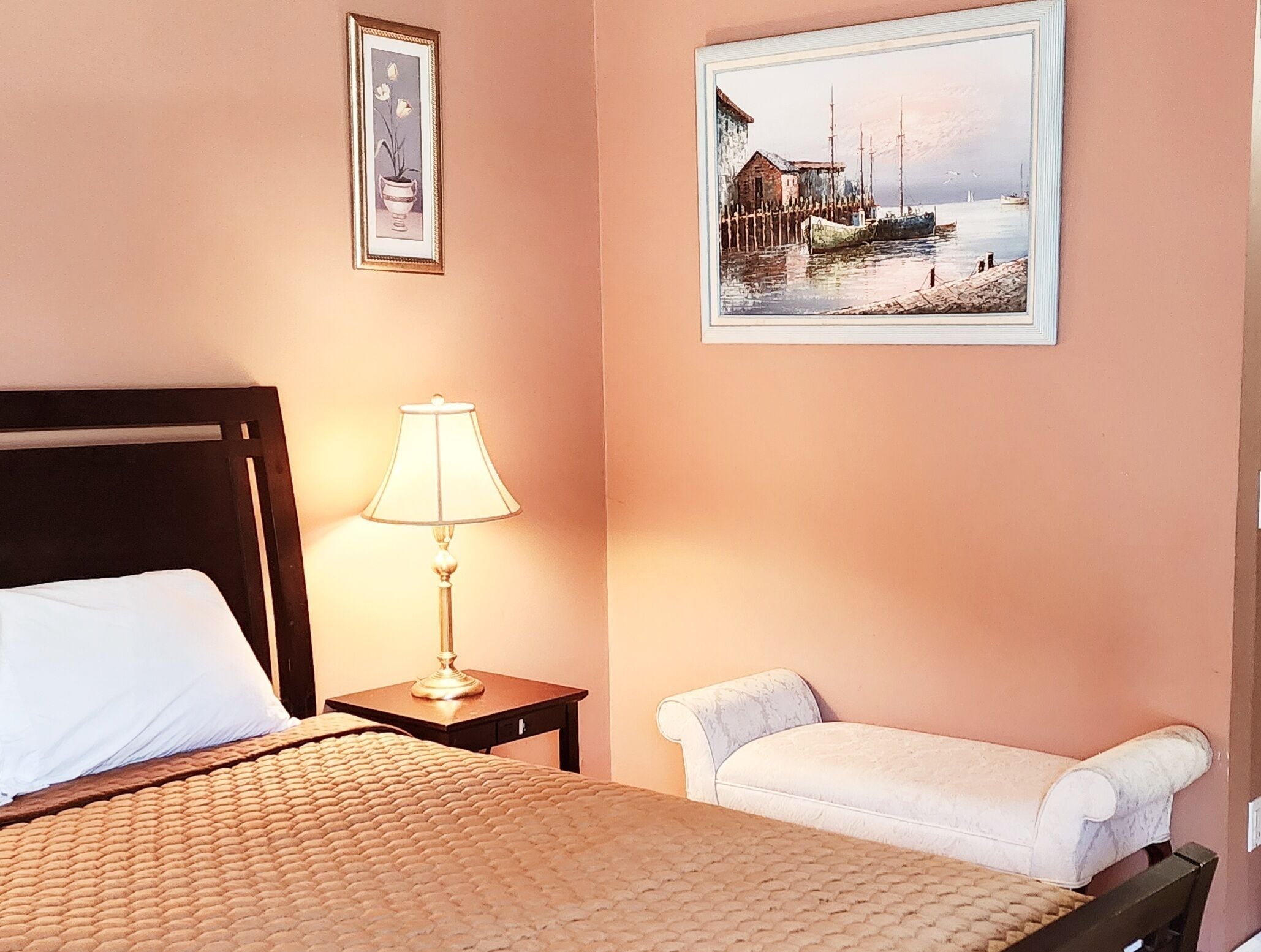 Une chambre d’hôtel avec un lit, une table de chevet avec une lampe, une chaise et deux tableaux encadrés au mur.