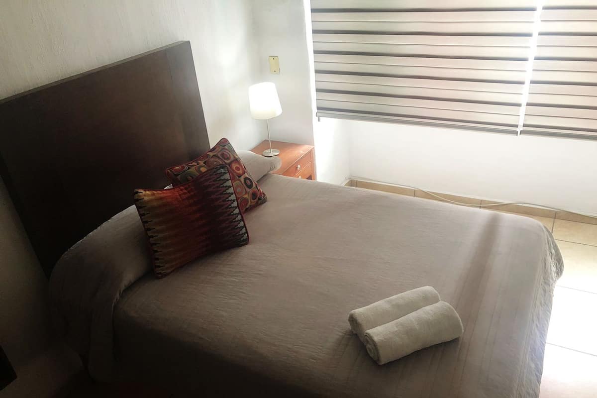 A neatly made bed with a folded towel, a patterned pillow, and a bedside lamp.