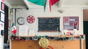 A reception desk with a 'h. HOSTELLIT' sign, a clock, a 'CHECKIN' sign, and a menu board. - Hostel Lit (Guadalajara)