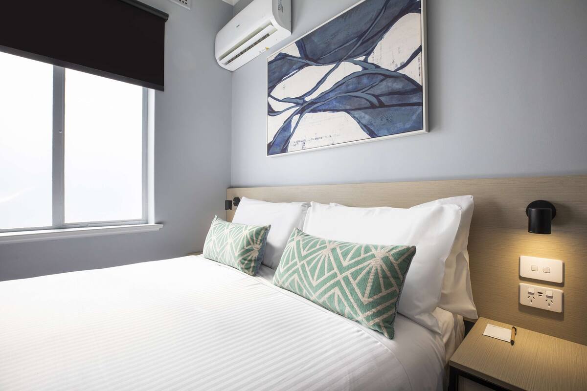 A hotel room with a large bed, a wall-mounted air conditioner, a painting, and a nightstand with a lamp and a power outlet.