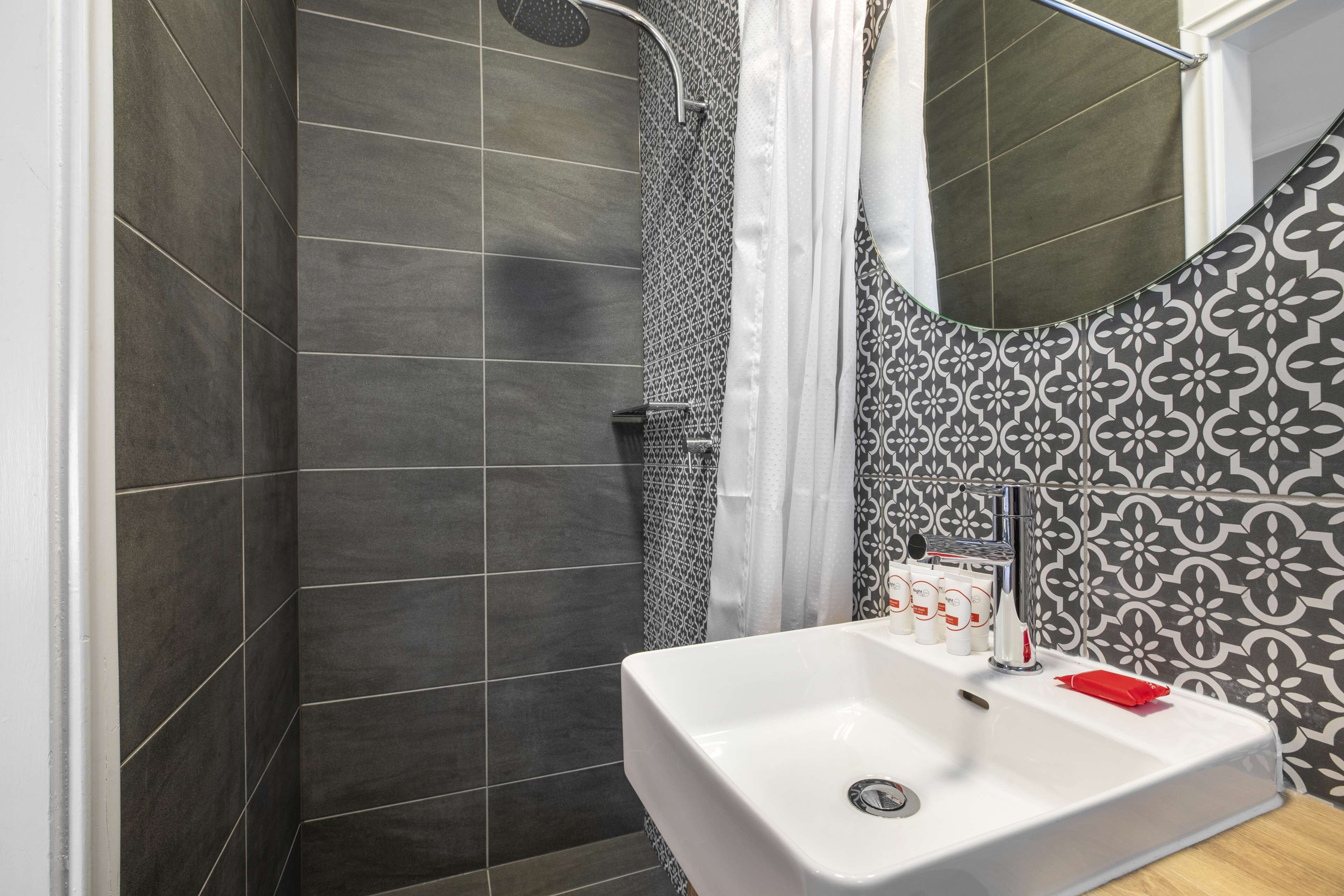 Un baño con paredes de azulejos oscuros, un lavamanos blanco con dispensador de jabón rojo, y una zona de ducha con un salpicadero de azulejos decorativos.