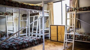 A dormitory room with bunk beds, wooden flooring, and a window with curtains.