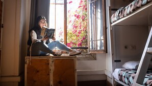 A person sitting on a wooden box in a room with a bunk bed, a window with a view of pink flowers, and a door. - Hostal Providencia - Hostel (Santiago)