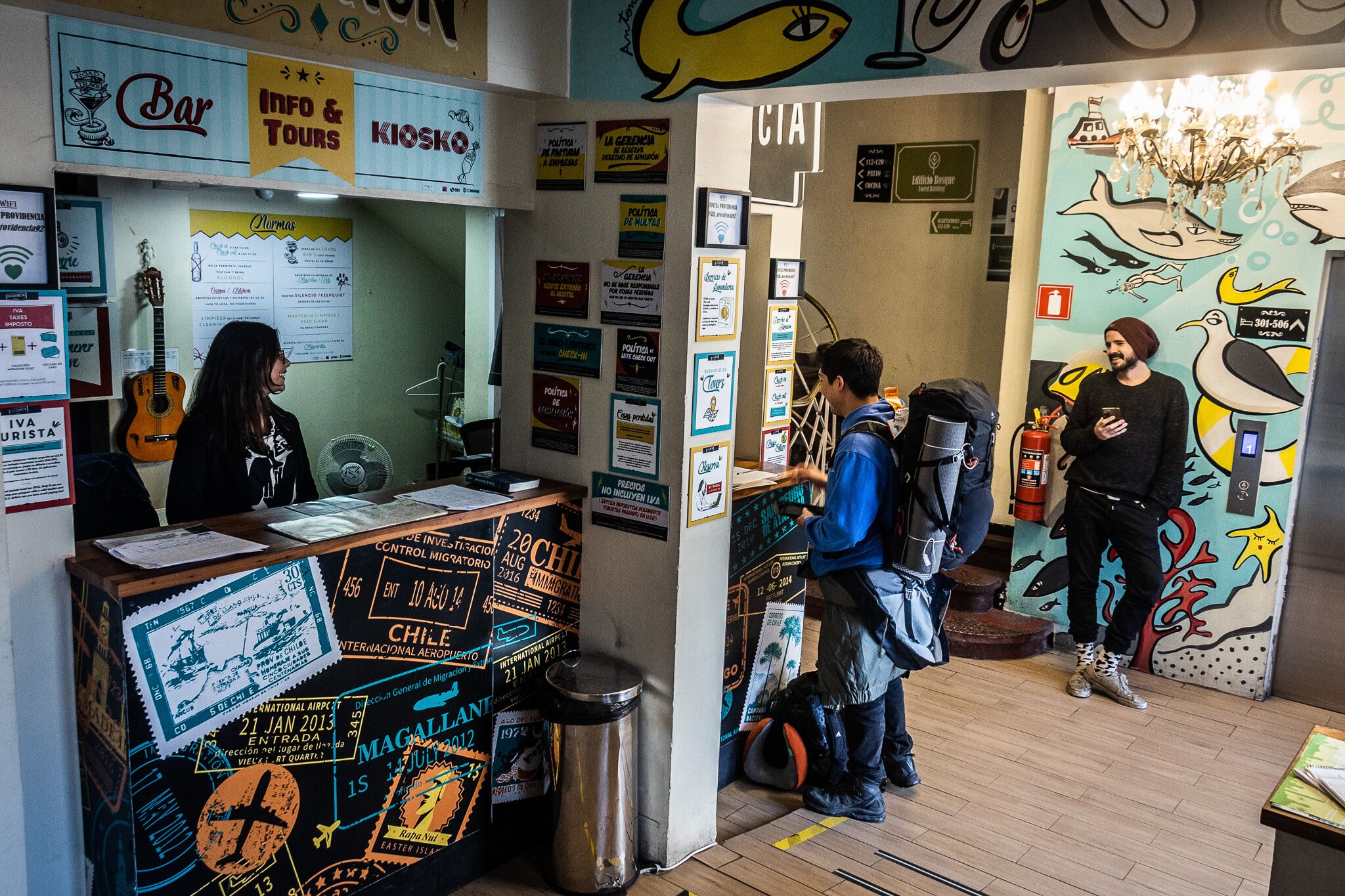 A bar area with a counter, posters, and a guitar. There are people interacting near the bar and a mural with birds on the wall.