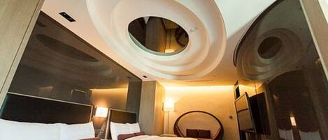 A modern hotel room with a circular skylight, a bed with white linens, a round seating area, and a large mirror on the wall.