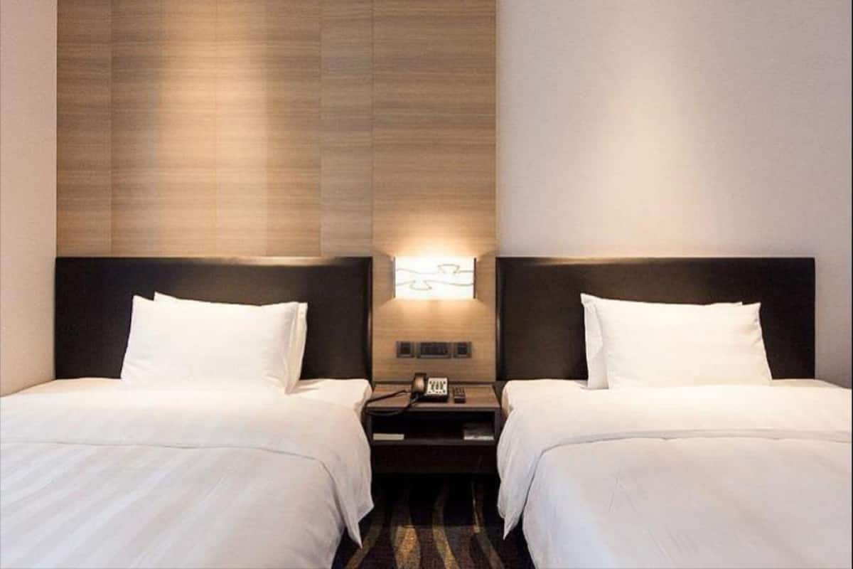 A hotel room with two beds, a nightstand between them, and a wall-mounted light fixture.