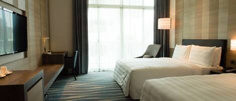 A hotel room with two beds, a desk, a TV, and a large window with curtains.