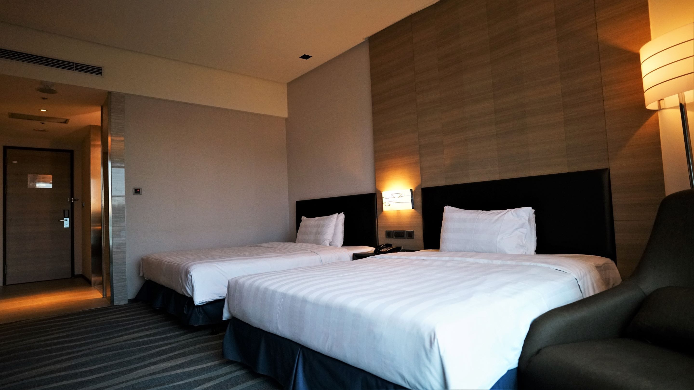 A hotel room with two beds, a desk, a chair, and a lamp.