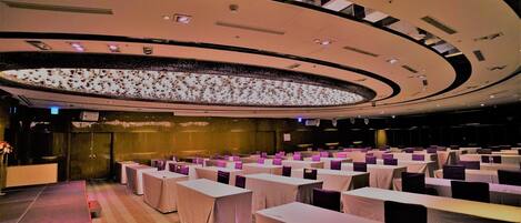 A conference room with rows of tables and chairs, a large circular ceiling feature, and a stage area.