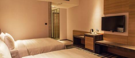 Habitación de hotel con dos camas, un escritorio, una silla, un televisor montado en la pared y un baño visible a través de una puerta de vidrio.