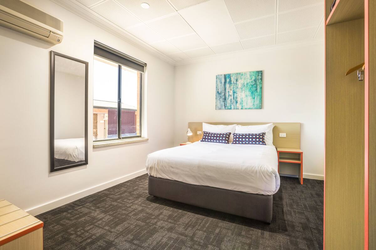 Ein Hotelzimmer mit einem großen Bett, einem Fenster, einem Spiegel, einem Bild und einem kleinen Nachttisch.