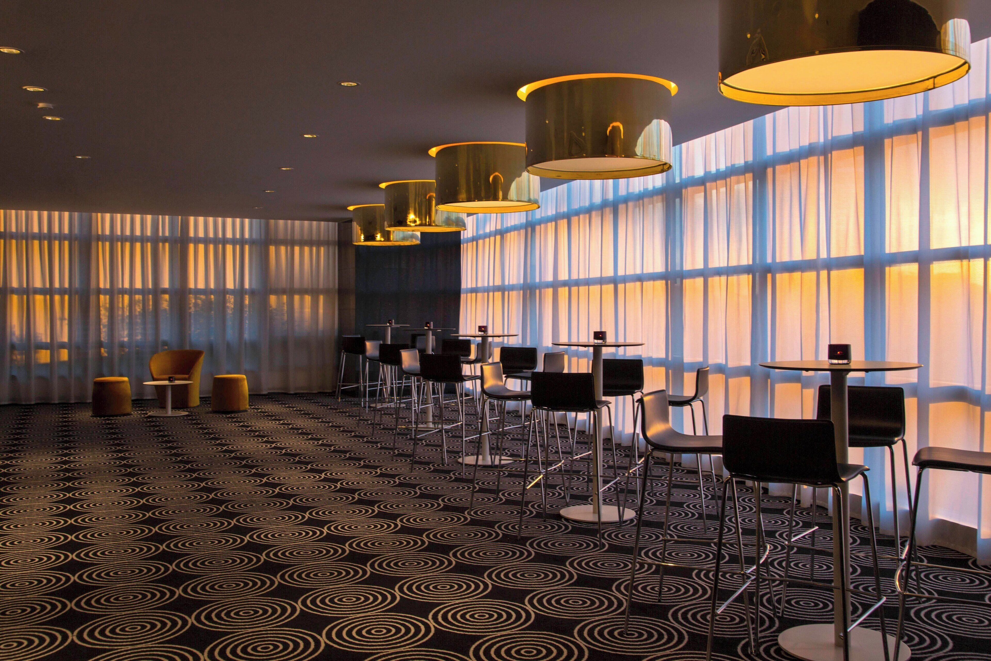 Un hall d’hôtel doté d’un tapis à motifs, de fauteuils et de tables modernes, ainsi que de grandes fenêtres ornées de rideaux.