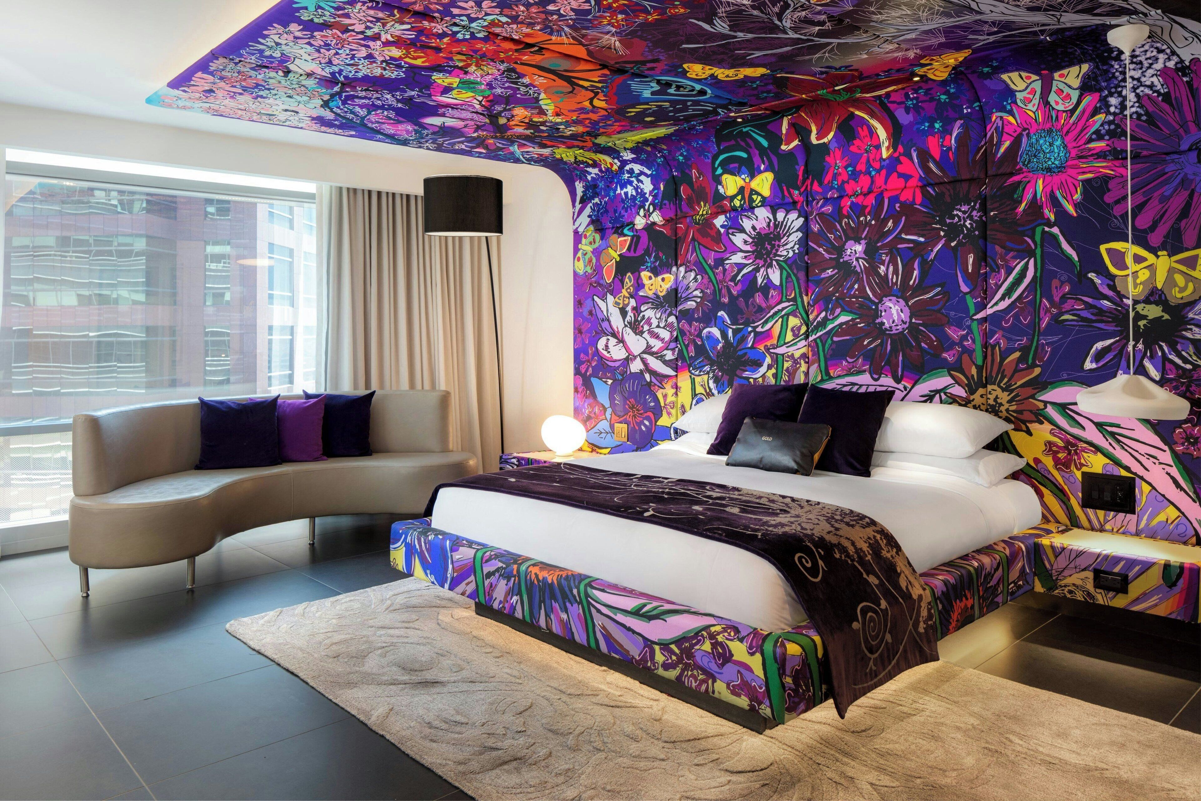A modern bedroom with a curved sofa, a large bed with a colorful floral headboard, and a vibrant mural on the wall.