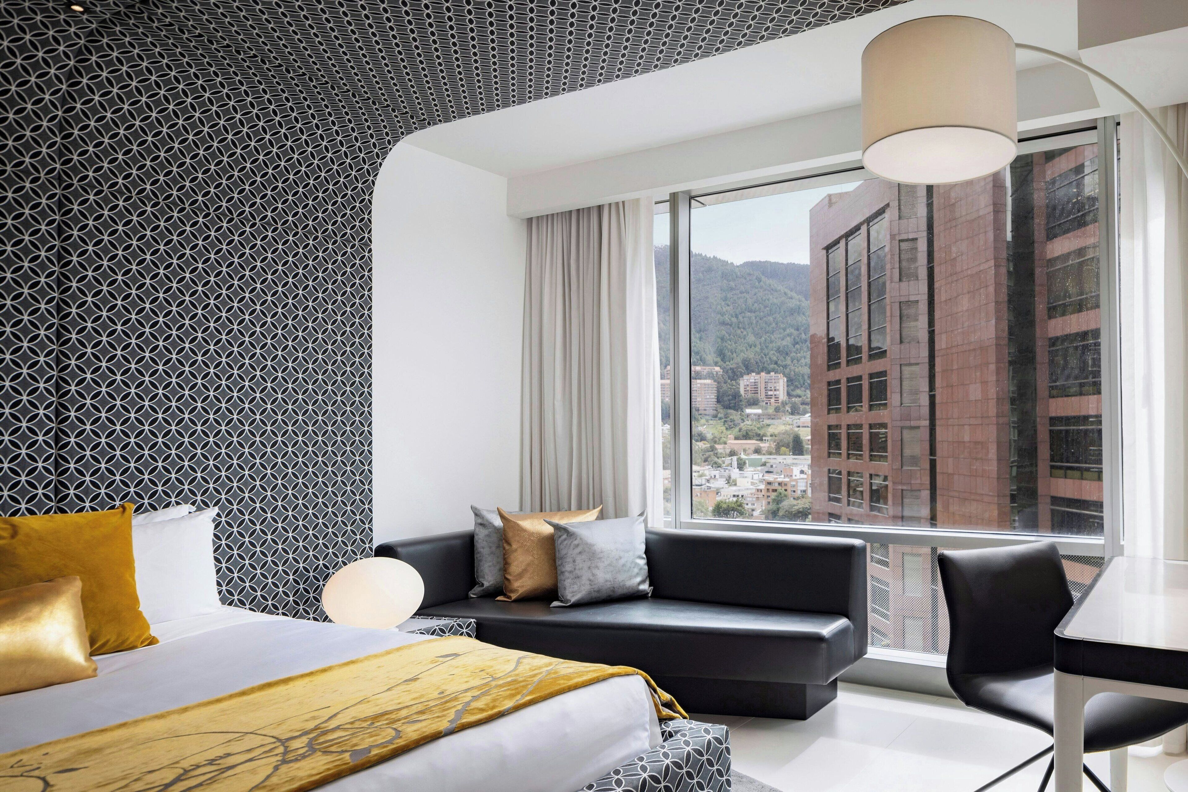 A modern hotel room with a large bed, a sofa, a desk, and a view of a cityscape through large windows.