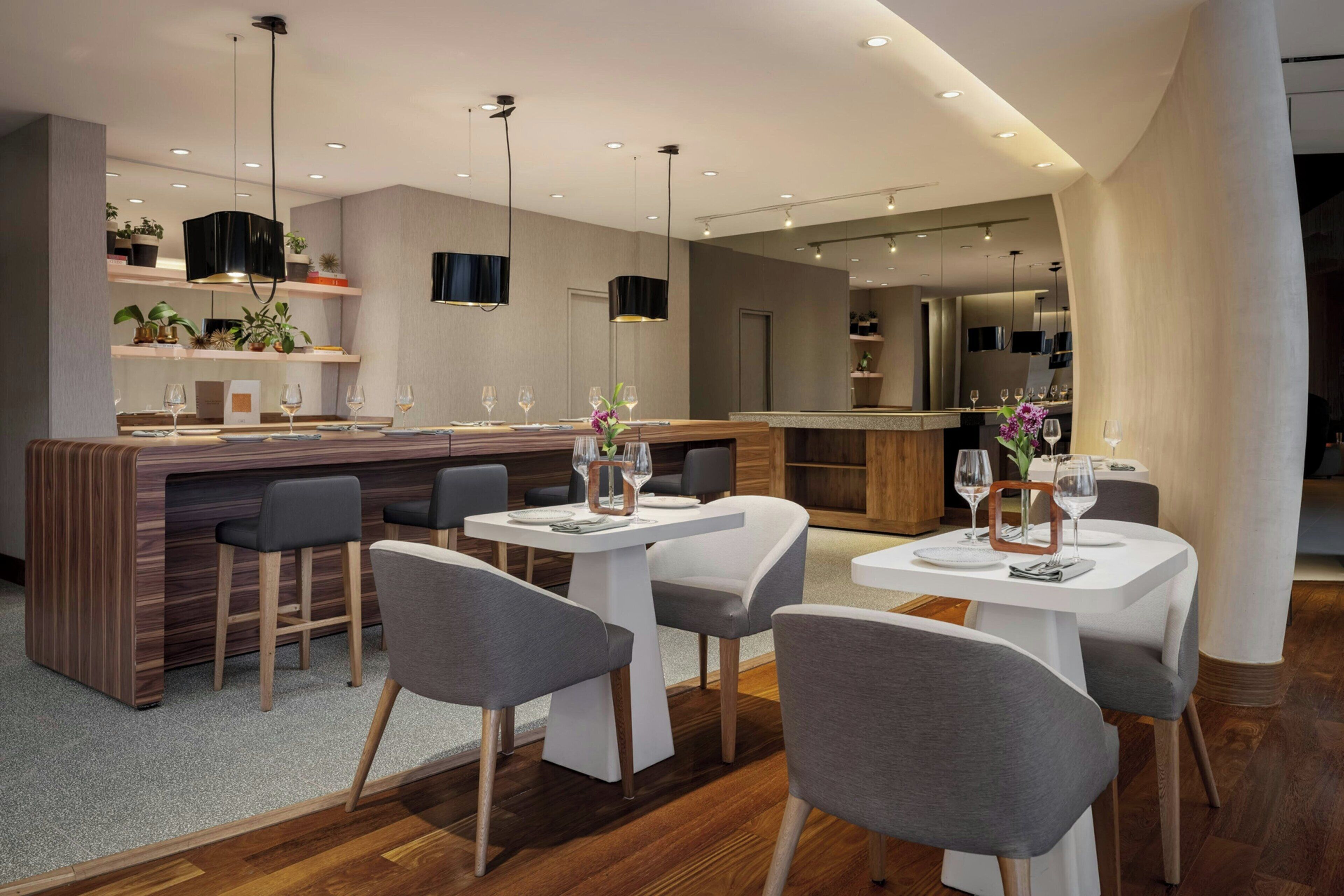Un espace de restauration moderne avec un bar, des tables blanches, des chaises grises et des luminaires suspendus.