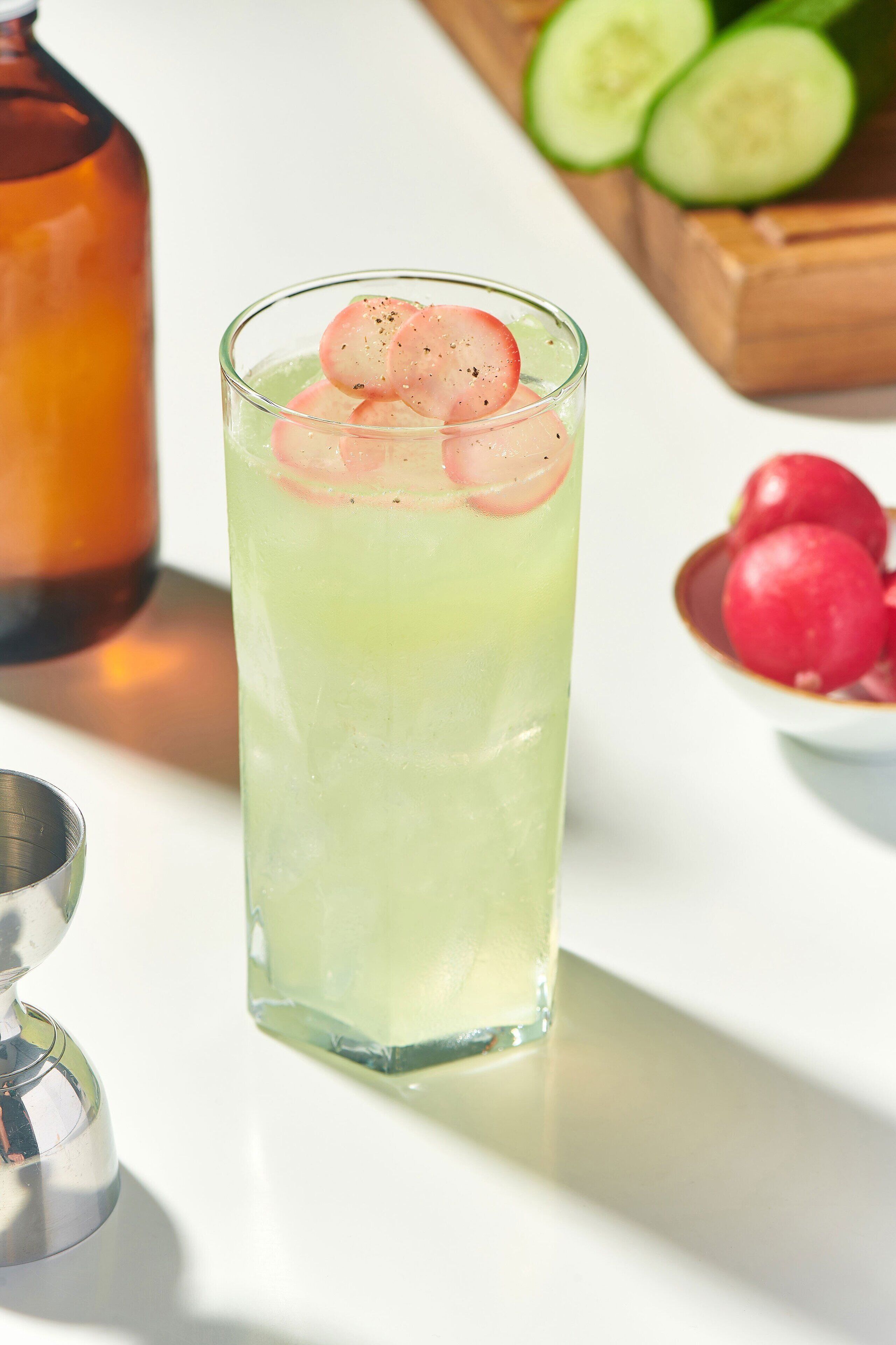 A tall glass with a green beverage, garnished with slices of radish and cucumber, on a white surface.