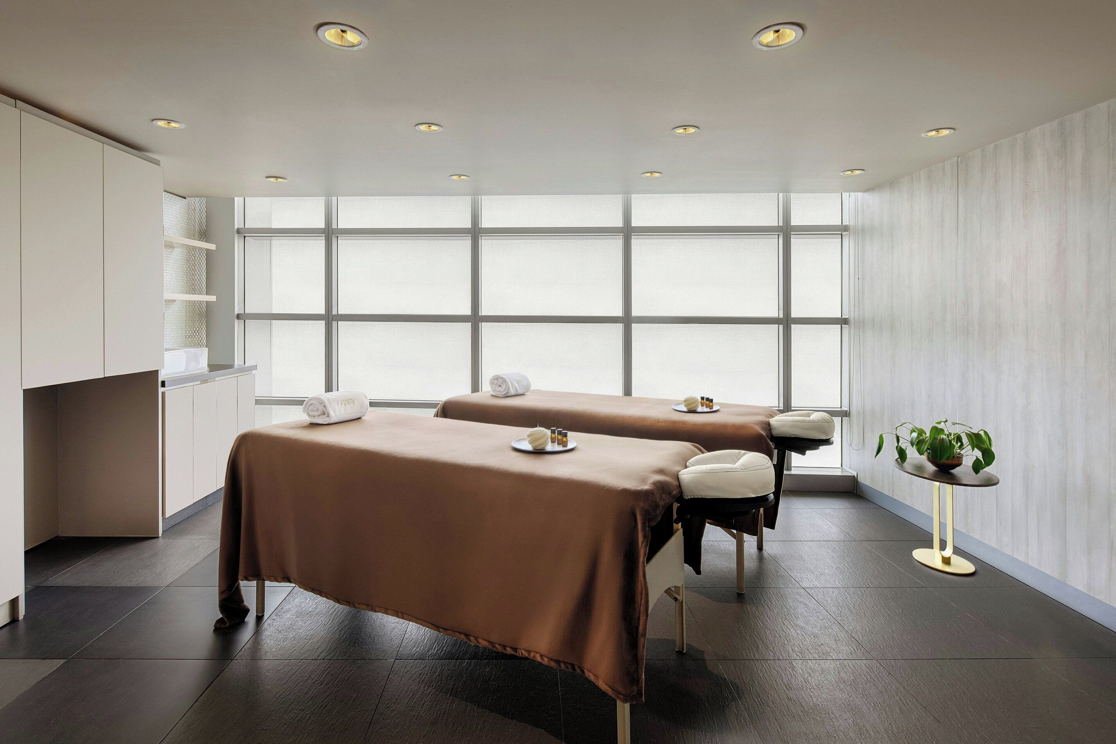 A spa room with two massage tables, a small round table with a plant, and a large window providing natural light.