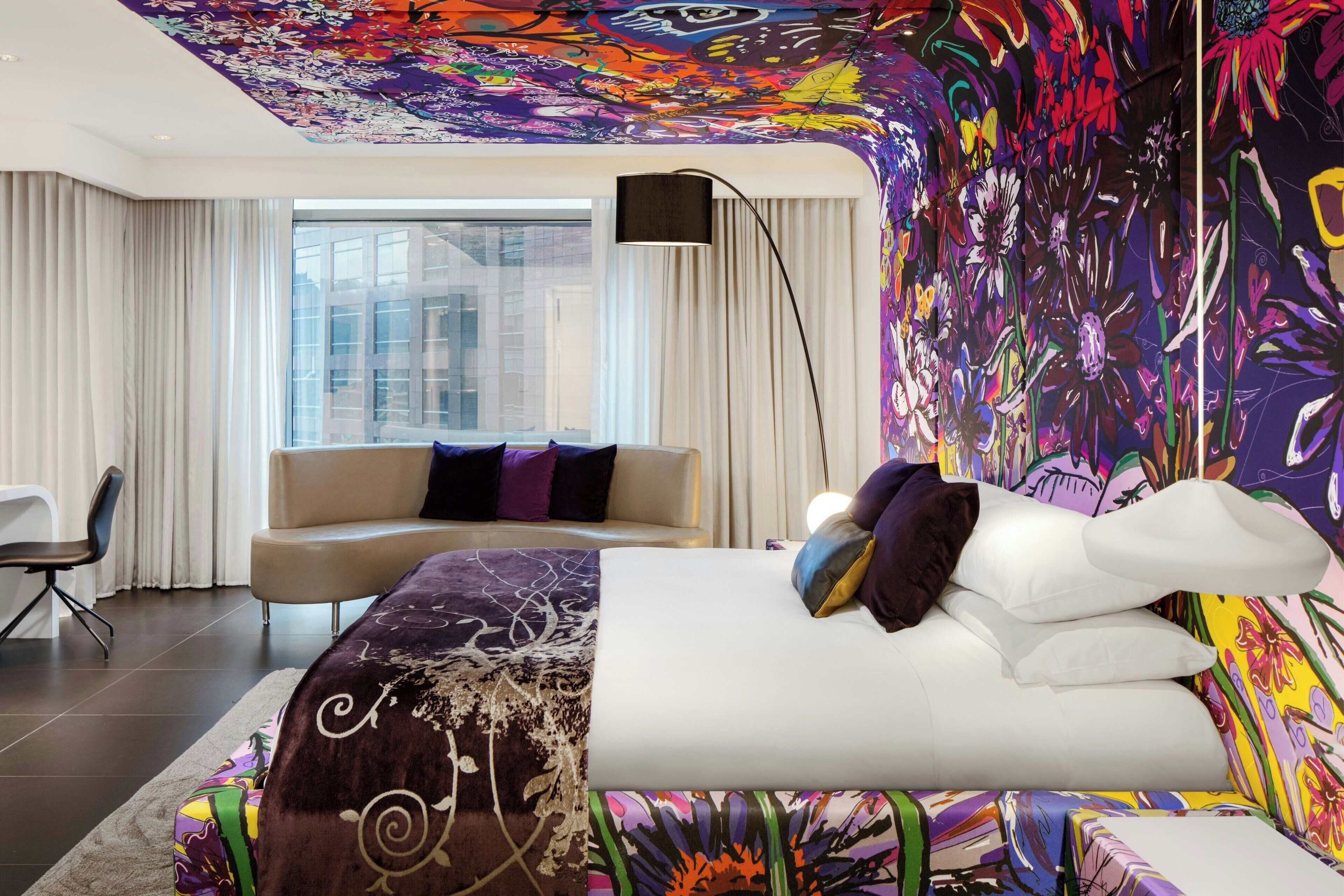 A modern hotel room with a large bed, a sofa, and a colorful abstract mural on the wall.