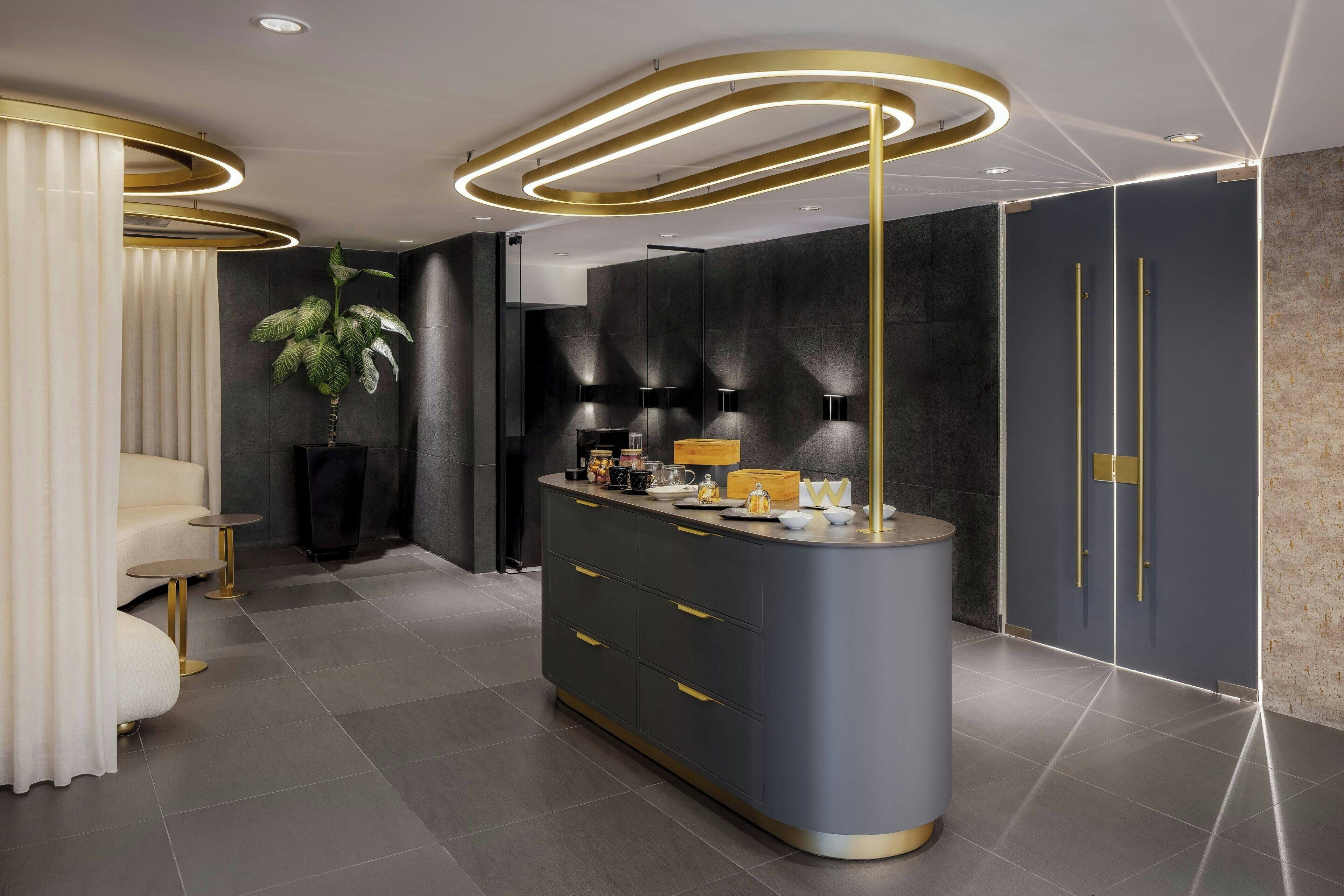 Un hall d’hôtel moderne avec un comptoir d’accueil incurvé, une plante en pot et un espace salon agrémenté d’un canapé blanc et de touches dorées.