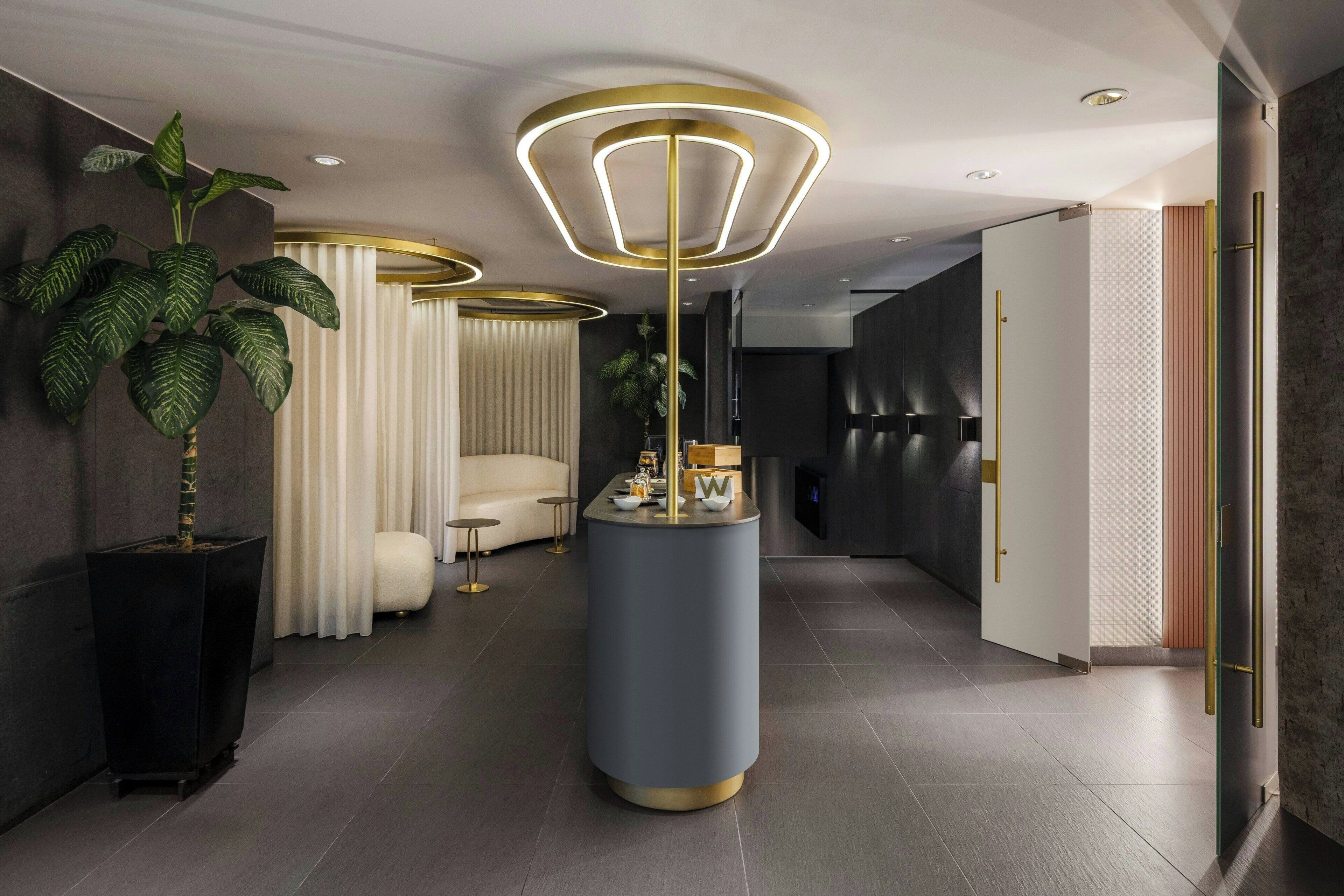 Un moderno vestíbulo de hotel con una recepción central, una planta grande en maceta, un sofá y un elemento decorativo de iluminación.