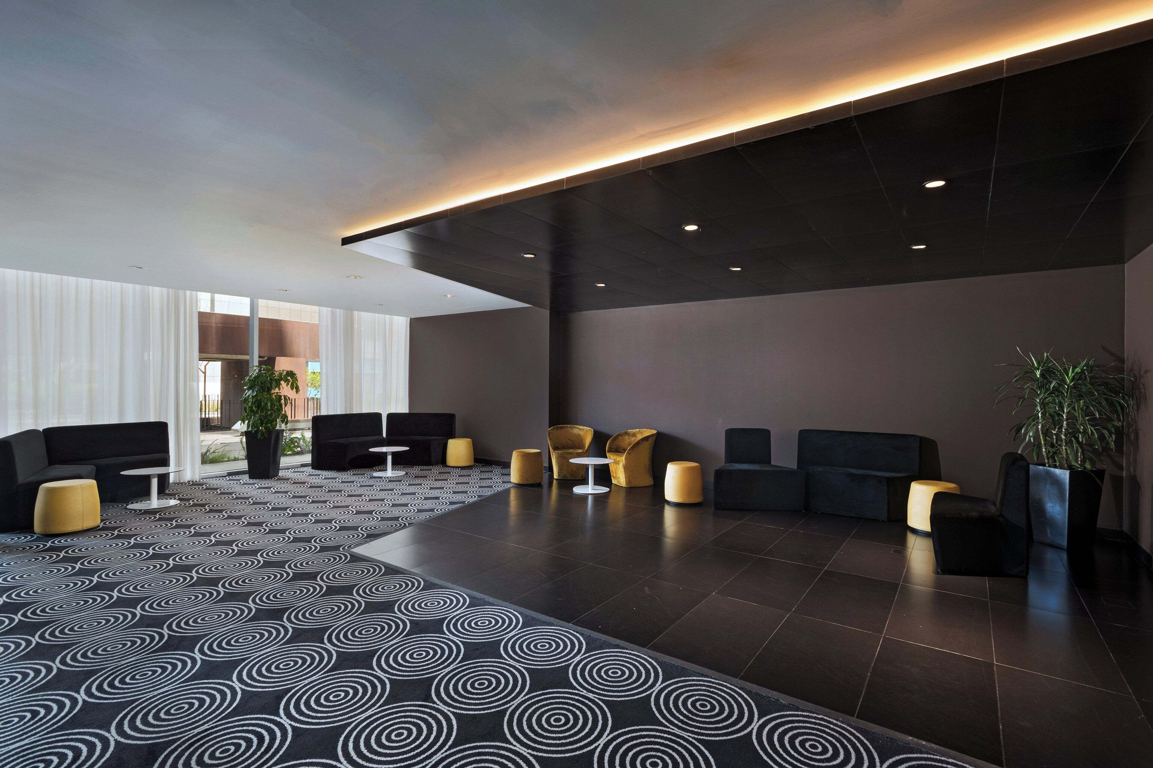 A modern lobby with a patterned carpet, black and gold seating, and a large potted plant.