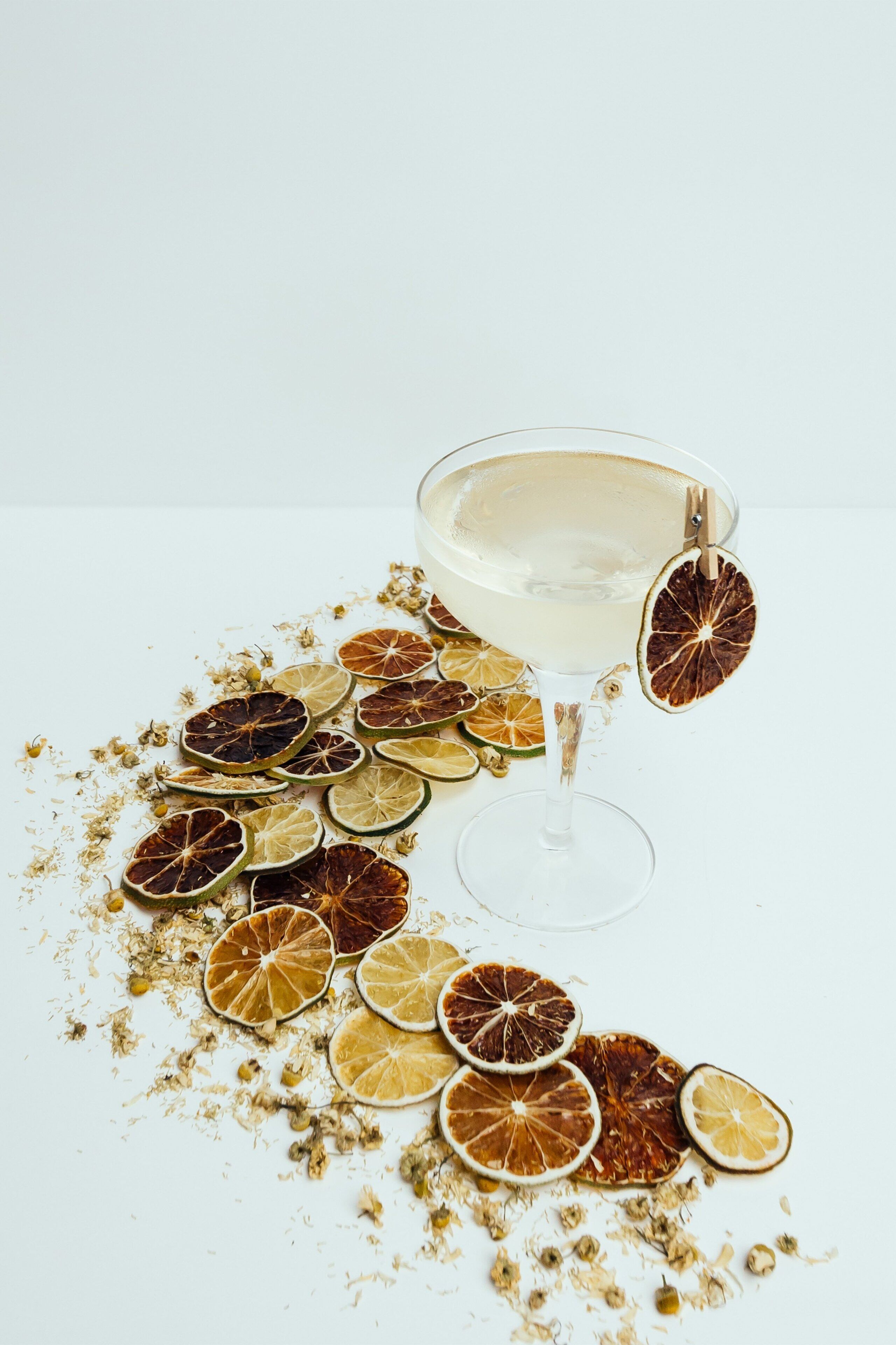 A clear glass with a drink on a white surface, surrounded by dried citrus slices and herbs.
