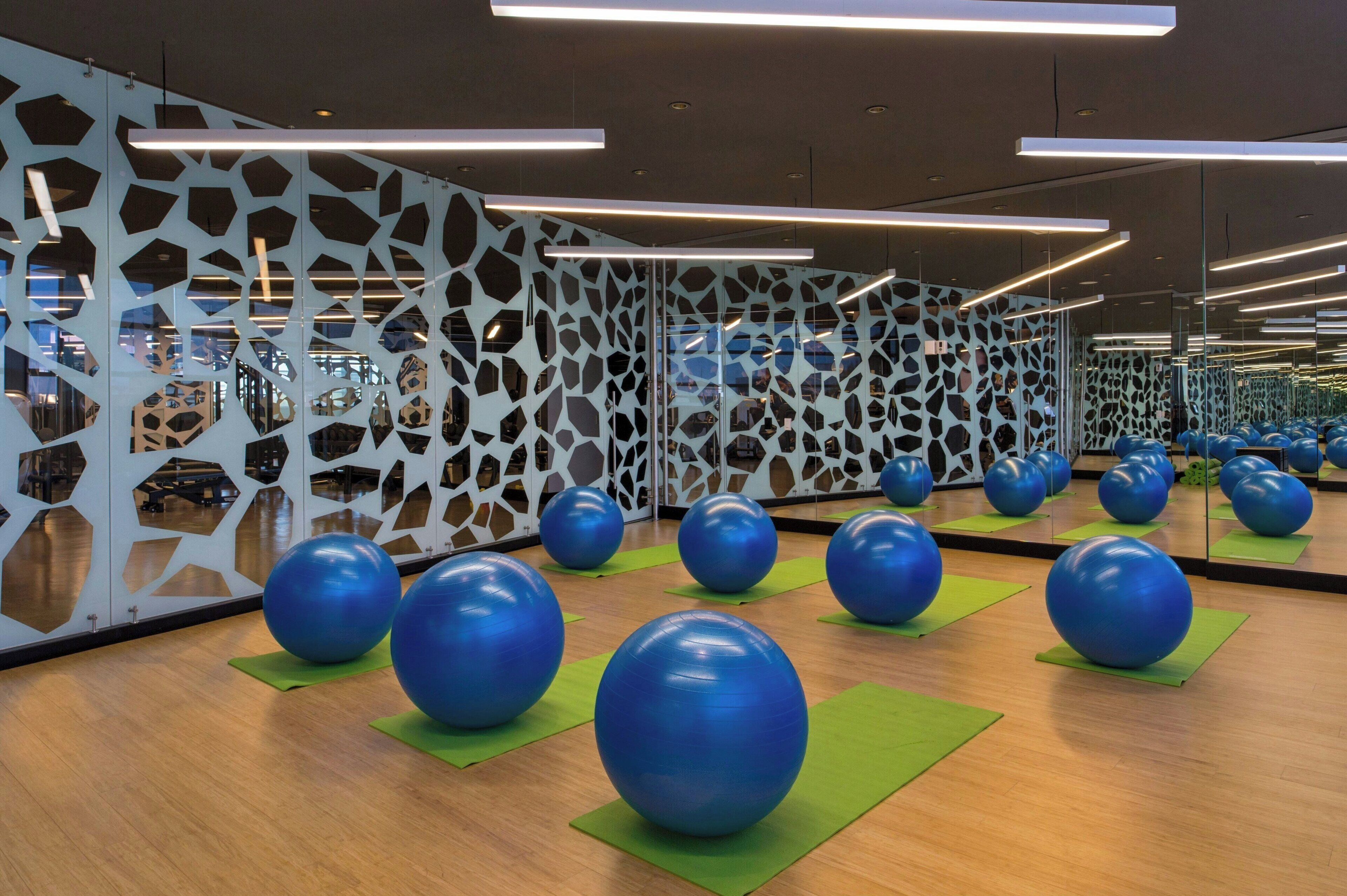Une salle de sport avec des ballons d’exercice sur des tapis verts, un plancher en bois et un mur orné de motifs géométriques découpés.