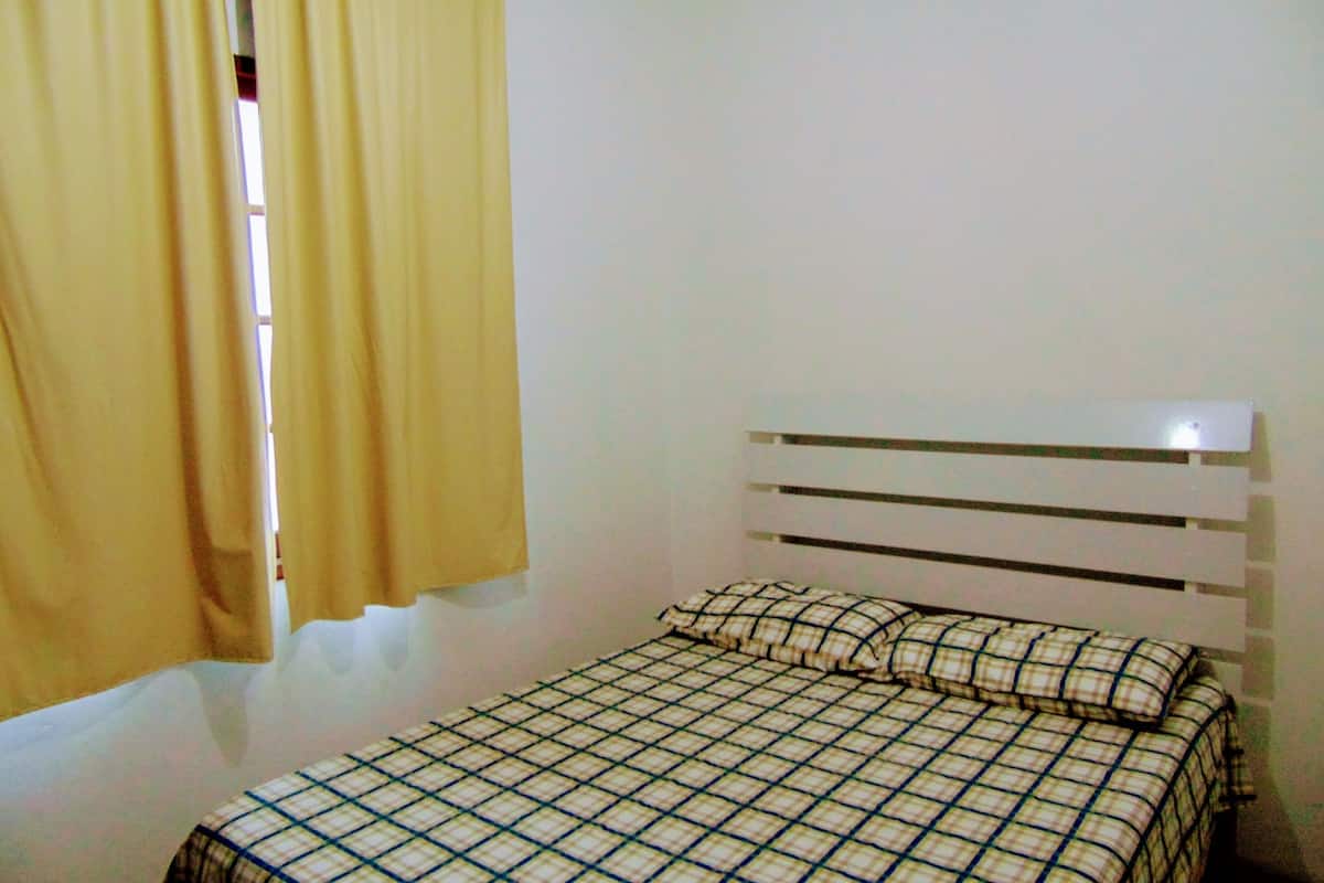 A small, simple room with a bed, a window with curtains, and a plain wall.