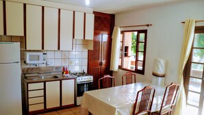 A compact kitchen with a stove, microwave, and refrigerator. There is a dining table with chairs and a window with curtains. - Ingleses Residence (Florianópolis)
