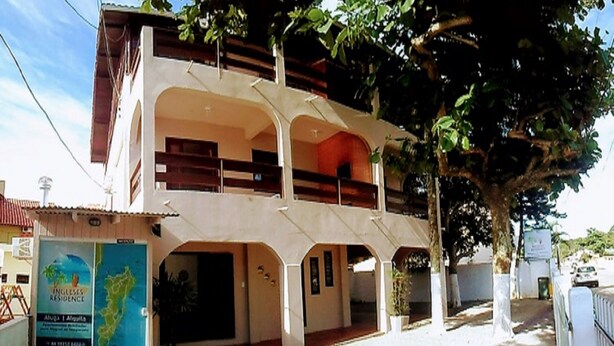 A two-story building with a balcony on the upper floor, a signboard advertising a residence, and a tree in the foreground. - Ingleses Residence (Florianópolis)
