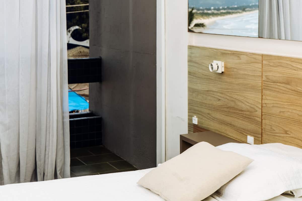 A bedroom with a bed, a wooden dresser, a mirror, and a view of the beach through a window.